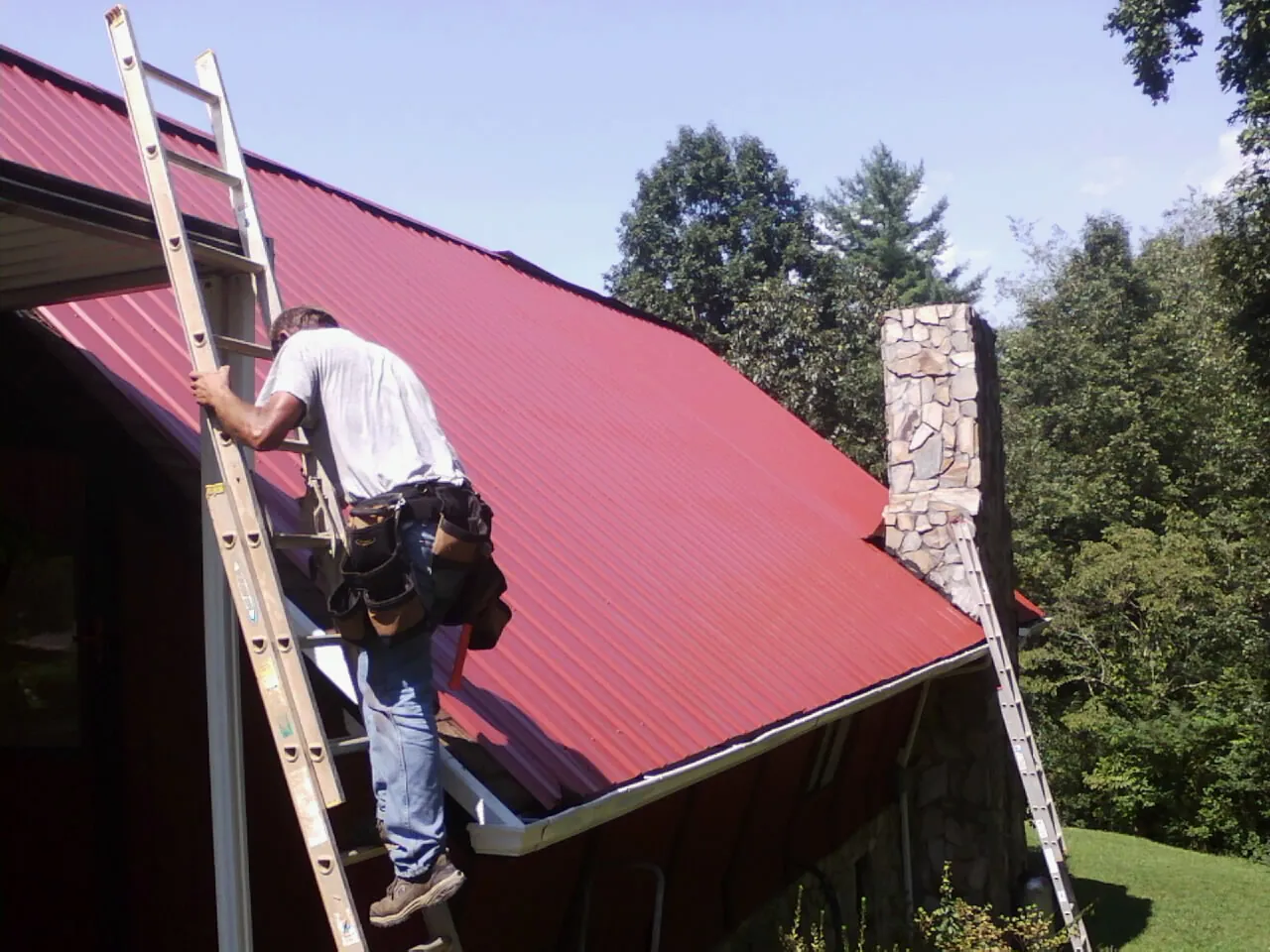 Roofer on a ladder beside a red metal roof and stone chimney — Soffit & Fascia Repair by Roofing Brothers in Glen Richey